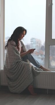 beautiful woman wrapped in a blanket sitting on the windowsill and using phone