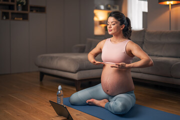 A relaxed pregnant woman doing breathing exercises at home while sitting on an exercise mat in front of a laptop