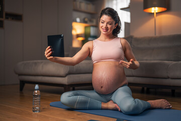 A healthy pregnant woman making an exercise video on a tablet while sitting on an exercise mat at home