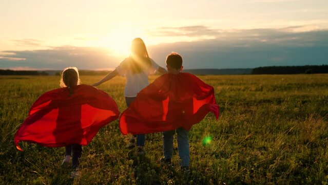 Mother daughter, son play superheroes in park. Mom child run in red raincoats, have fun outdoor. Family Carnival in sun, Halloween, sun. Mom, Super kids dream of becoming superhero, flying in red cape