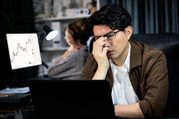 Businessman with stressful face working on laptop waiting email while coworker trading stock...