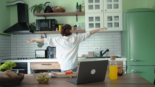 A young Caucasian woman is having fun and dancing while cooking in a brightly lit kitchen.