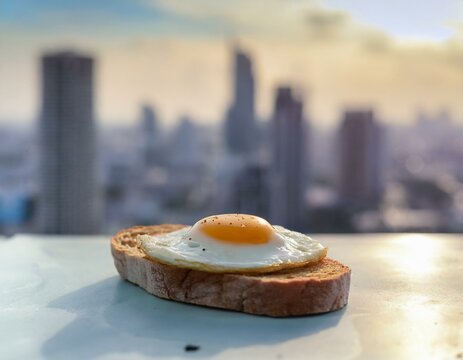 A fried egg atop a rustic bread slice, on a sleek, modern table on a city rooftop, with skyscrapers in the soft-focus background, blending urban life with the simplicity of breakfast.