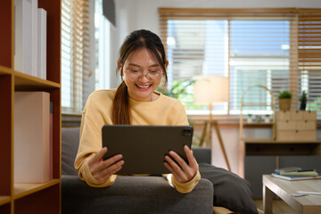 Happy teenage woman in glasses using digital tablet on couch at home