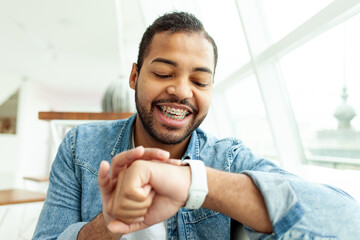 happy african american man with braces smiles and looks at his wristwatch in a white cafe, a man in...