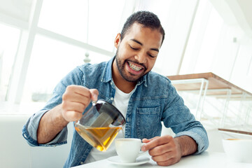 happy african american man with braces holds a teapot and pours tea into a cup in a white cafe, the...