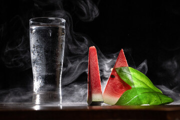 A glass of cold fresh water and watermelon with leaf on a black background