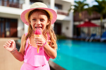 Smiling girl in pink swimsuit enjoys ice cream cone by poolside at summer resort. Child indulges in frozen treat on sunny day, cooling off near water. Happiness, leisure at vacation hotspot.