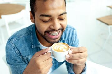 African American man with braces drinks coffee from a cup and smiles in a white room, a man with...