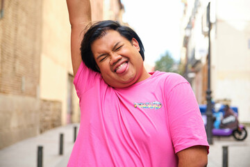 Gay Asian man in a pink shirt is holding her arm up and making a funny face. The shirt has the word...