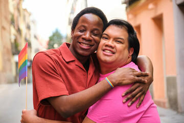 Gay multiracial couple hug each other while one of them holds a rainbow flag. Scene is happy and...