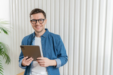 A poised man with glasses, holding a tablet, stands smiling in a bright room adorned with indoor greenery.