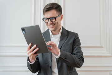 An enthusiastic businessman, clad in a smart grey suit, navigates a tablet with a pleased...