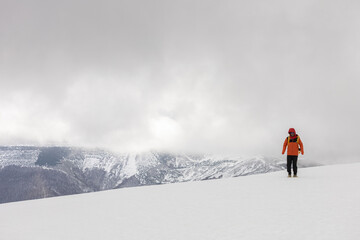 A man in an orange jacket is walking on a snowy mountain. The sky is cloudy and the mountains are...