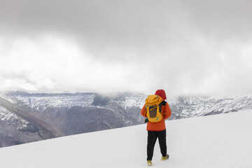 A person wearing a red jacket and a yellow backpack is standing on a snowy mountain. The sky is...