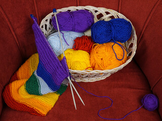Colorful balls of wool for knitting and hand-knitted scarf in rainbow colors lie in a wicker basket standing in a red velour armchair
