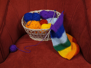 Colorful balls of wool for knitting and hand-knitted scarf in rainbow colors lie in a wicker basket standing in a red velour armchair

