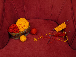 Skeins of red and orange wool in wicker basket, balls of red and orange wool and samples knitting pattern lie on a red velour armchair
 