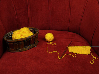 Skein of yellow wool in wicker basket, ball of yellow wool and sample knitting pattern lie on a red velour armchair
 