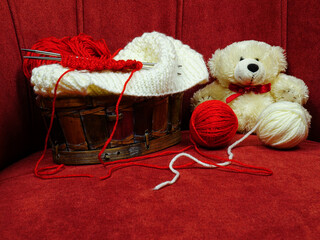A wicker basket with accessories for hand knitting and a white teddy bear are in the chair