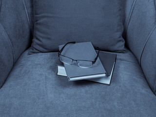 There are glasses, closed books and a dark gray decorative pillow in a gray velour armchair 
