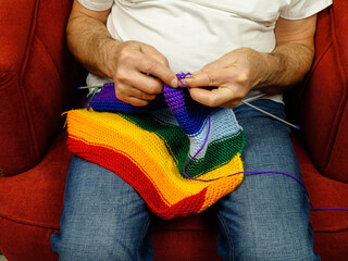 Person, sitting in an armchair, knits a scarf in rainbow colors on knitting needles 