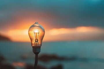 An incandescent light bulb perfectly aligns with the setting sun, creating an illusion of being lit by the sunlight against a blurred ocean background. - obrazy, fototapety, plakaty