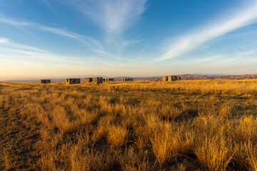 Evening view of an earthen road through the savannah and an abandoned village. Bright blue sky with clouds, orange grass and unfinished houses. Low mountains around. Georgia.