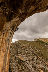 A person is climbing a rock wall with a view of a mountain in the background. The sky is cloudy, and the mountain is covered in trees