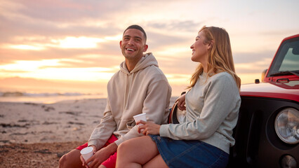 Young Couple Standing Chatting By Car With Hot Drink At Beach Watching Sunrise Together