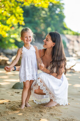 Young woman mother with a little daughter in white dresses on seashore in the shade of trees and palms