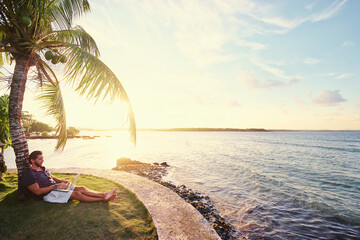 Work and vacation. Young man working on laptop computer on the tropical beach under the palm tree.