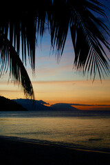 Sunset on a tropical island. View of the sea and the silhouette of a hilly tropical island through palm branches.
