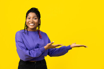 A young woman presenting with an open hand gesture in a purple sweater against a vibrant yellow...