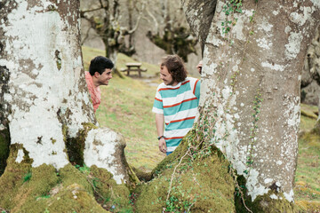 A playful and happy male couple hide and seek around the trunks of majestic, moss-covered trees, exuding a sense of joy and togetherness in a serene forest