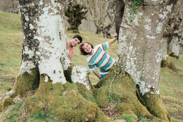 A playful and happy male couple hide and seek around the trunks of majestic, moss-covered trees, exuding a sense of joy and togetherness in a serene forest