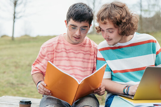 Two men, engaged in their studies, share an orange folder full of notes while sitting at a park bench, a laptop open before them