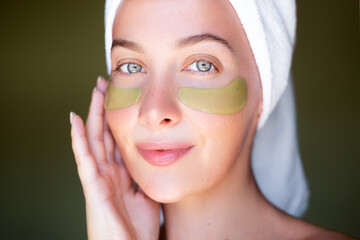A serene woman with a towel on her head and green hydrogel eye patches smiles softly against a dark...