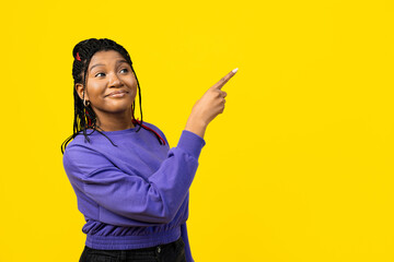 A cheerful woman pointing to the side and looking away isolated on a vibrant yellow background,...