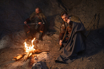 Medieval knights sharpen swords by fire in cave - 773976636