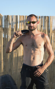 Shirtless european man with sunglasses near a wooden fence at dusk. Stronger then yesterday. Young handsome man in sportswear holding tank top and looking at camera. Strong man with a beard.