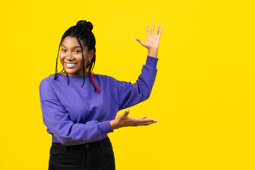 Afro woman presenting something on a vivid yellow background, conveying positivity and enthusiasm. - obrazy, fototapety, plakaty