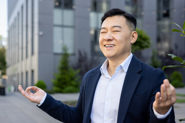 Close-up photo of young Asian male businessman sitting relaxed outside working office in lotus yoga pose and meditating with closed eyes