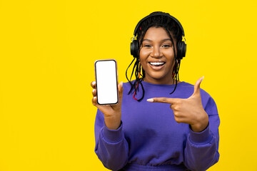 Woman pointing at a blank smartphone screen, wearing in headphones and purple sweater, ideal for...