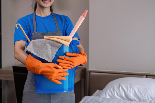 The cleaning lady wears orange rubber gloves and carries a bucket of bedroom cleaning supplies.