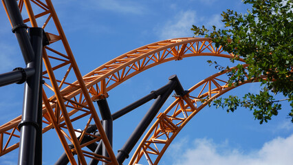 Orange-coloured roller coaster tracks of an extreme roller coaster, crossing under a blue sky and a...