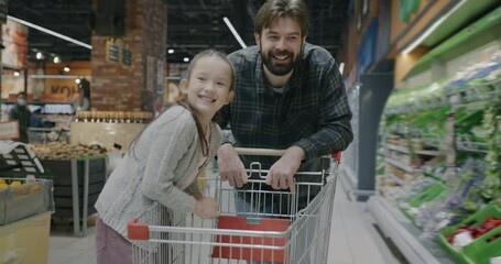 Slow motion portrait of young man father pushing shopping cart with little girl daughter in supermarket while kid laughing having fun. Family and shop customer concept. - Powered by Adobe