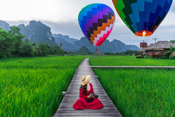 Asian woman holding a camera sits on a wooden walkway and looks at balloons with green rice fields in Vang Vieng, Laos