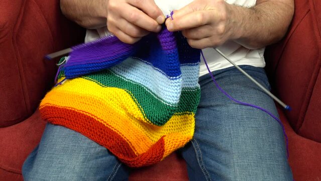 Person, sitting in an armchair, knits a scarf in rainbow colors on knitting needles 