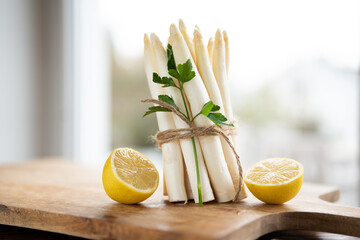 Standing bunch of fresh white asparagus. Seasonal spring vegetables with lemon and flat-leaf parsley on wooden cutting board. Kitchen scene for the seasonal gastronomy. - 774022086